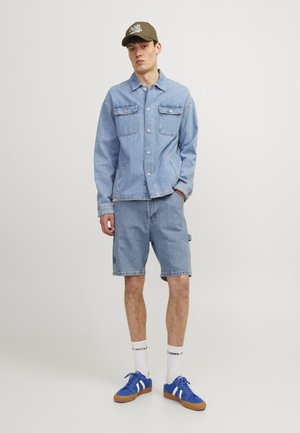 Chaqueta de mezclilla azul claro con dos bolsillos en el pecho, combinada con shorts de mezclilla azul claro. Zapatillas azules con rayas blancas y calcetines blancos.