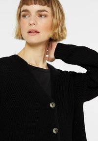 Schwarze gestrickte Strickjacke mit V-Ausschnitt und langen Ärmeln. Verfügt über große, runde Knöpfe und ein strukturiertes Muster. Geeignet zum Überziehen.