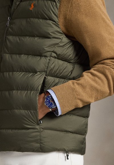 Olivengrønn puffervest med quiltet tekstur, båret over en brun genser. En blå klokke med rustfritt stålarmbånd titter frem fra mansjetten.