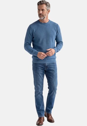 Uomo con capelli grigi e barba che indossa un maglione blu, jeans blu e scarpe di pelle marrone, in piedi davanti a uno sfondo bianco.