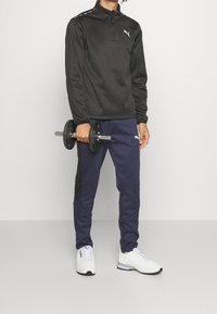 Černá sportovní mikina na zip, námořnické kalhoty s černými bočními panely, držící činky. Bílé tenisky s síťovinou a gumovými akcenty.