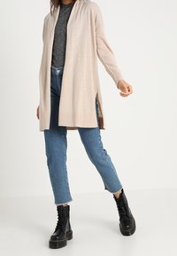 Mujer vistiendo un cárdigan largo beige, una camiseta gris oscuro, jeans cortos azules y botas de plataforma negras con cordones, de pie con una pierna doblada.