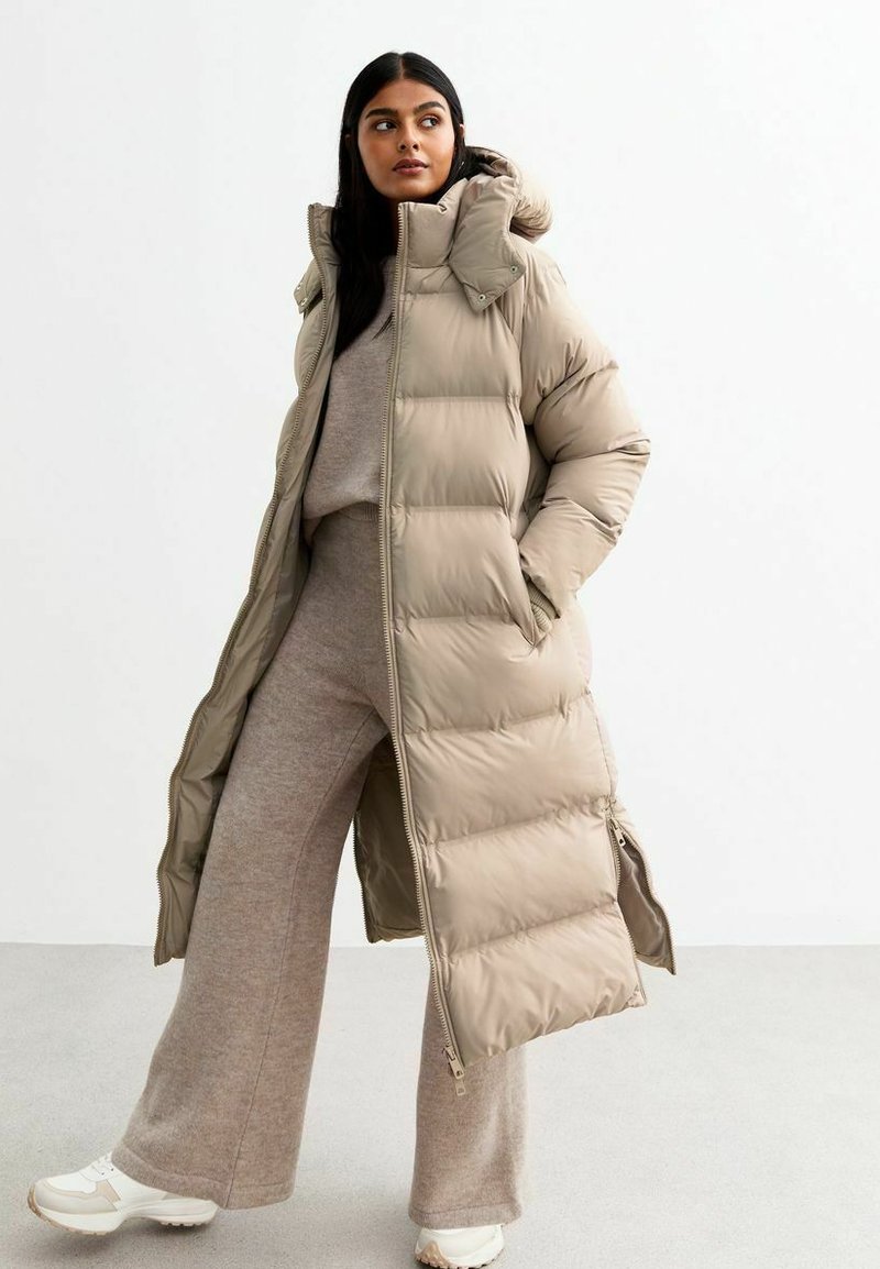 Długi beżowy puchowy płaszcz z kapturem, z delikatnym pikowaniem, zamek błyskawiczny i boczne kieszenie. Nosi się go na jasnoszarym stroju.