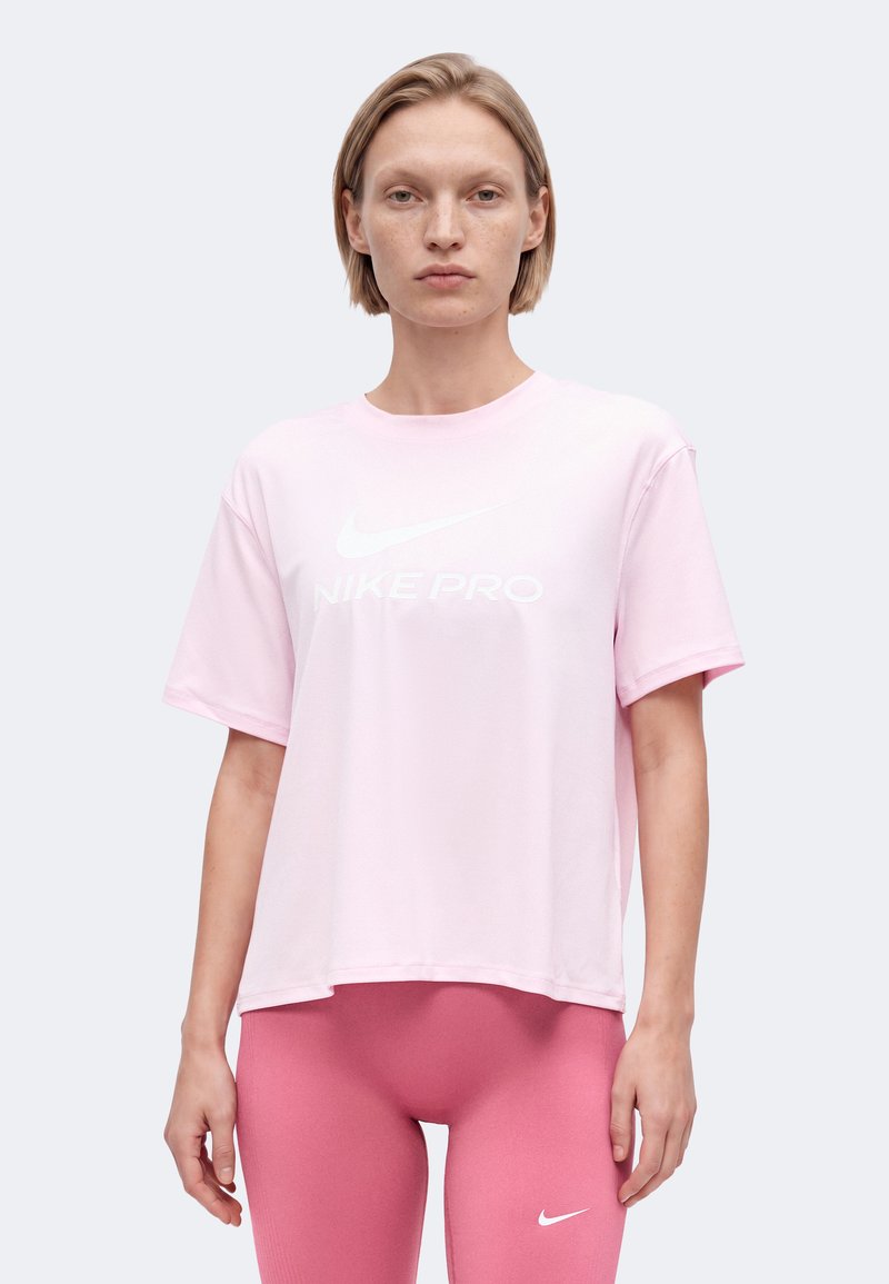 Γυναίκα που φοράει ροζ Nike Pro t-shirt σε ανοιχτή απόχρωση και πιο σκούρο ροζ κολάν με λογότυπα Nike, στέκεται μπροστά σε απλό ανοιχτό γκρι φόντο.