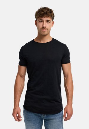 Młody mężczyzna z kręconymi włosami, ubrany w dopasowany czarny T-shirt z krótkim rękawem i niebieskie dżinsy, stoi na tle jednolitego białego tła.