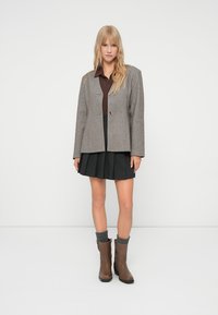 Blazer gris con un solo botón, combinado con una camisa marrón de canalé y una falda negra con pliegues. Botines marrones con puños grises completan el look.