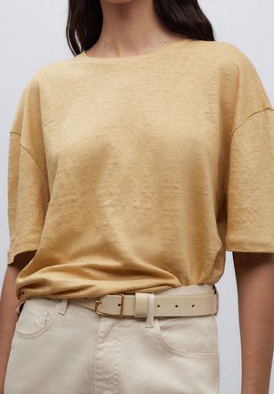 Donna che indossa una blusa corta gialla senape, larga, infilata in pantaloni a vita alta di colore bianco sporco, con una cintura beige.