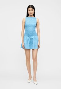 Γυναίκα που στέκεται φορώντας ένα αμάνικο, ανοιχτό μπλε υφασμάτινο μίνι φόρεμα και λευκά μυτερά τακούνια, μπροστά σε λευκό απλό φόντο.