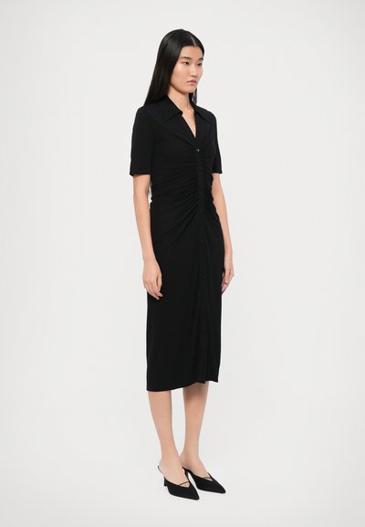 Robe noire à boutons avec un col, des manches courtes et un design froncé. Porte avec des chaussures noires à talons bas et à bout pointu.