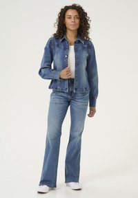 Denim jakna z zapenjanjem na gumb, s ostrim ovratnikom in zahodnimi yoki, v kombinaciji s širokimi kavbojkami. Oboje v srednji modri pranjeni barvi. Bele superge.