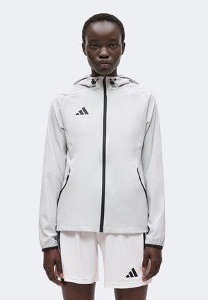 Kvinde iført en lysegrå hættetrøje med sort lynlås og hvide shorts, begge med sort Adidas-logo.