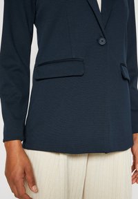 Blazer bleu marine avec deux poches et un bouton. Fabriqué à partir d'un tissu texturé, il présente un revers cranté et une silhouette ajustée, associé à un pantalon clair.