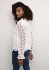 White lace blouse with a geometric pattern and ruffled sleeve cuffs, paired with blue jeans. Features sheer, textured details.