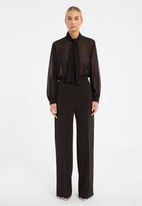 Blouse noire transparente avec un nœud cou, associée à un pantalon noir taille haute à jambes larges présentant des paillettes discrètes. Les deux pièces ont des manches longues.
