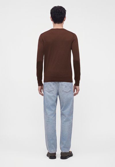 Homme debout de dos, portant une chemise marron à manches longues, un jean bleu clair et des chaussures foncées sur un fond uni.