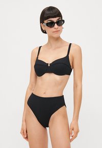 Čierny rebrovaný bikiny s balcónovým topom, ktorý má nastaviteľné ramienka a zapínanie na prednej strane. Vysoký pás s miernym zakrytím.