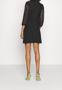 Vestido corto negro con mangas abullonadas de encaje transparente. Combinado con sandalias de tacón alto verdes con tiras, resaltando las piernas y detalles del tobillo.