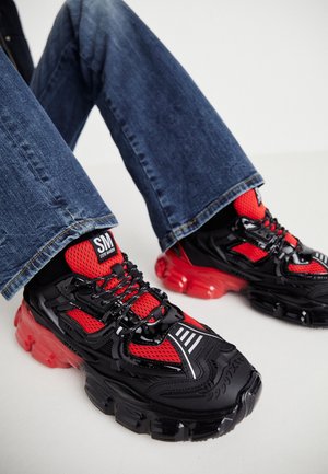 Persoon draagt een blauwe spijkerbroek en zwart-rode chunky sneakers met het label "SM Steve Madden" en zit met gebogen benen.