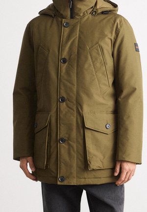 Hombre con abrigo de invierno marrón con capucha, botones delanteros, bolsillos con solapa y bolsillos en el pecho en ángulo, combinado con pantalones oscuros.