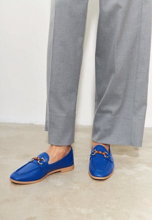 Personne portant un pantalon gris à jambes larges et des mocassins en cuir bleu vif avec semelles couleur cannelle et détail en métal, debout sur un sol clair.
