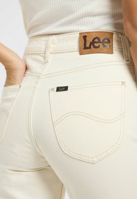 Los pantalones vaqueros de color crema presentan una etiqueta de cuero marrón con "Lee" en la cintura y una pequeña etiqueta negra en el bolsillo trasero.