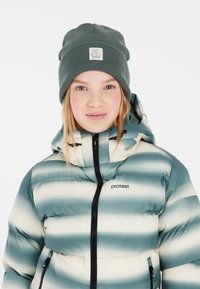 Puffjacke mit blau-grünen und weißen horizontalen Streifen, schwarzem Reißverschluss und Logo auf der linken Brust. Strickmütze in Grün mit Patch-Logo.