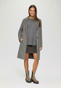 Manteau en laine gris avec un col cranté, associé à un pull rayé gris et une jupe sombre, complété par des bottines gris avec des accents noirs.