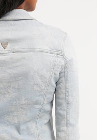 Giacca in denim azzurro chiaro con sottili motivi floreali indossata da una persona, mostrando il retro e un piccolo logo metallico triangolare vicino al colletto.