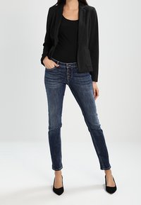 Blazer negro en una tela texturizada, combinado con una blusa negra ajustada y jeans skinny de mezclilla azul. Modelo llevando tacones negros de punta.