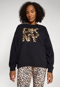 Zwarte sweatshirt met gedurfde luipaardprint "DKNY" logo. Combinatie met beige legging met een bruine luipaardprint. Krullend haar zichtbaar.