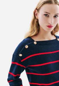 Pull bleu marine avec des rayures horizontales rouges, doté de boutons dorés à l'épaule et d'un col bateau. Texture en maille douce.