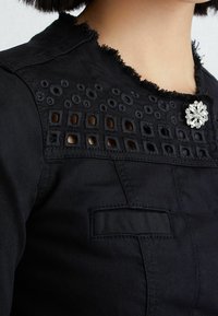 Dettaglio di un indumento nero con ricami a intaglio quadrato e circolare vicino al collo, bordi sfrangiati e una spilla decorativa con strass.