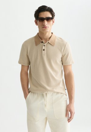 Man draagt een bruin polo met patroon, lichtbeige broek met trekkoord en donkere zonnebril, staand met handen in de zakken.
