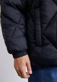 Juodos puffer tipo striukės rankovė su geometriniu siuvinėjimu, elastinga rankove ir šoniniu kišeniu. Matoma ranka laiko audinį.