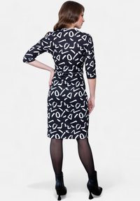 Robe noire avec des motifs abstraits blancs, manches trois-quarts et silhouette ajustée, accompagnée de bottines. Texture satinée.