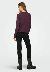 Pull en maille violet foncé avec un ajustement décontracté, doté d'un motif décoratif sur les épaules, associé à un jean skinny noir et des bottes noires à talons.