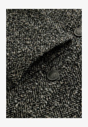 Tessuto in misto lana con un motivo houndstooth tessurato, prevalentemente nero e beige, con un dettaglio in metallo scuro sul bottone.
