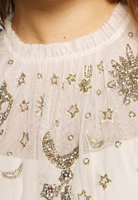 Genomskinlig krämfärgad tyg med guld- och klara kristallsbroderier som föreställer stjärnor och halvmånar, samlad halsringning och texturerade detaljer.