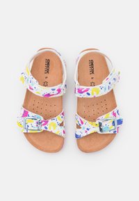 Värikärpäset sandaalit valkoisella yläosalla, jossa on abstrakteja vaaleanpunaisia, keltaisia ja sinisiä kuvioita. Korkki pohja ja hopea solkiyksityiskohta.