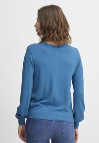 Pulover tricotat albastru cu mâneci lungi, ușor bufante și gât rotund, asortat cu blugi din denim. Textură netedă și detalii cu tiv nervurat.