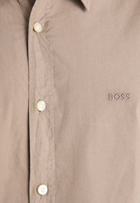 Camisa de botón marrón claro hecha de tela suave. Presenta un cuello puntiagudo, un logotipo en el pecho, botones crema y detalles cosidos.