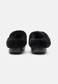 Fekete, bolyhos slip-on papucsok gumitalppal, hátulról, fehér háttér előtt látható.
