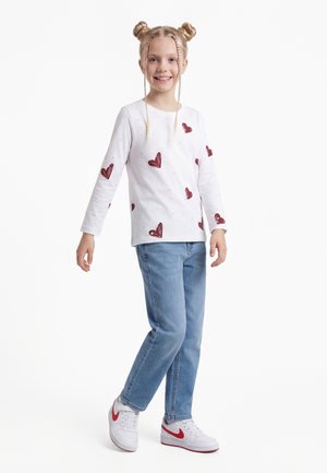 Lachend meisje met blonde dubbele knotjes, gekleed in een wit shirt met lange mouwen met rode hartjes, lichtblauwe spijkerbroek en witte sneakers met rode strepen.