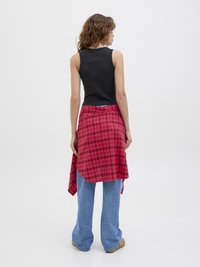 Top de malha preta canelada combinado com uma camisa de flanela vermelha em padrão xadrez, atada na cintura, usada com jeans de perna larga azul claros e calçado bege.