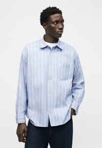 Άνδρας που φοράει πουκάμισο με λευκές και γαλάζιες ρίγες μανίκι μακρύ, πάνω από λευκό τοπ, με σκούρο παντελόνι, κοιτάζοντας στο πλάι σε μονόχρωμο φόντο.