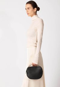 Zwarte leren handtas met een afgeronde vorm, bovenhandvat en ritssluiting, gedragen door een persoon die een lichtgekleurde top en rok draagt.