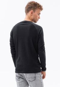 Svart långärmad tröja i strukturerad tyg, raglanärmar och rundad halsringning. Bakifrån, ihop med ljusgrå jeans och armband.