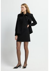 Schwarze Jacke mit mit Juwelen besetzten Taschen, getragen über einem schwarzen Kleid. Das Kleid hat eine strukturierte Passform und ist kombiniert mit schwarzen Slingback-Heels mit spitzen Zehen.