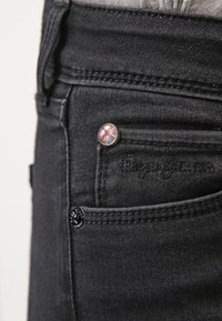Los jeans de mezclilla negros cuentan con un pequeño bolsillo frontal, herrajes en plata y negro, con un botón que muestra un diseño de la Union Jack.