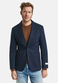 Marineblauer Woll-Blazer mit Revers, zwei Vordertaschen und sichtbarem Etikett. Trägt man über einem braunen Pullover und kombiniert mit hellen Jeans.